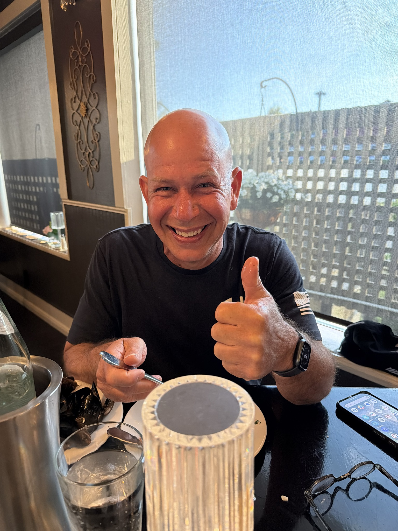 A smiling man with a shaved head gives a thumbs up while sitting at a restaurant table with water, utensils, and a large candle in the foreground. Sunlight streams through the window behind him.