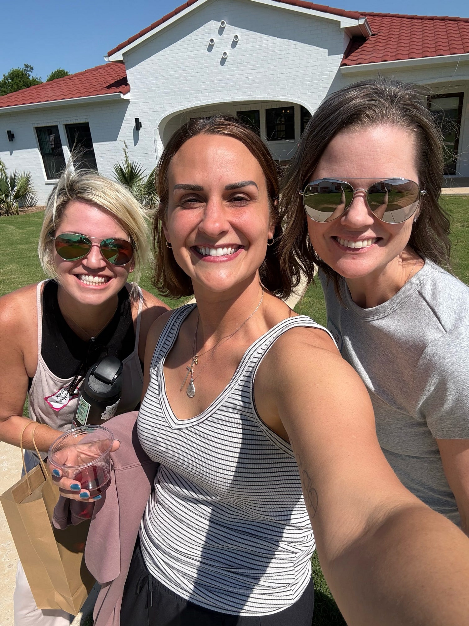Three smiling women take a selfie outside on a sunny day in front of a white house with a red roof. One woman holds a drink and another carries a bag. All are wearing sunglasses and casual clothes.