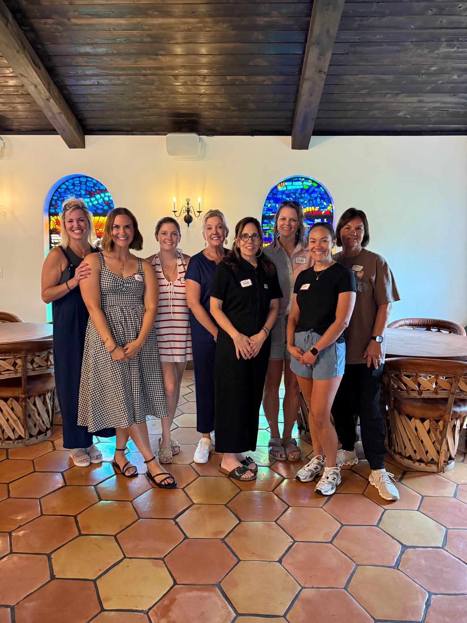 Eight women stand together and smile in a restaurant with colorful stained glass windows, wooden ceiling, and tiled floor. They wear casual summer clothing and name tags.
