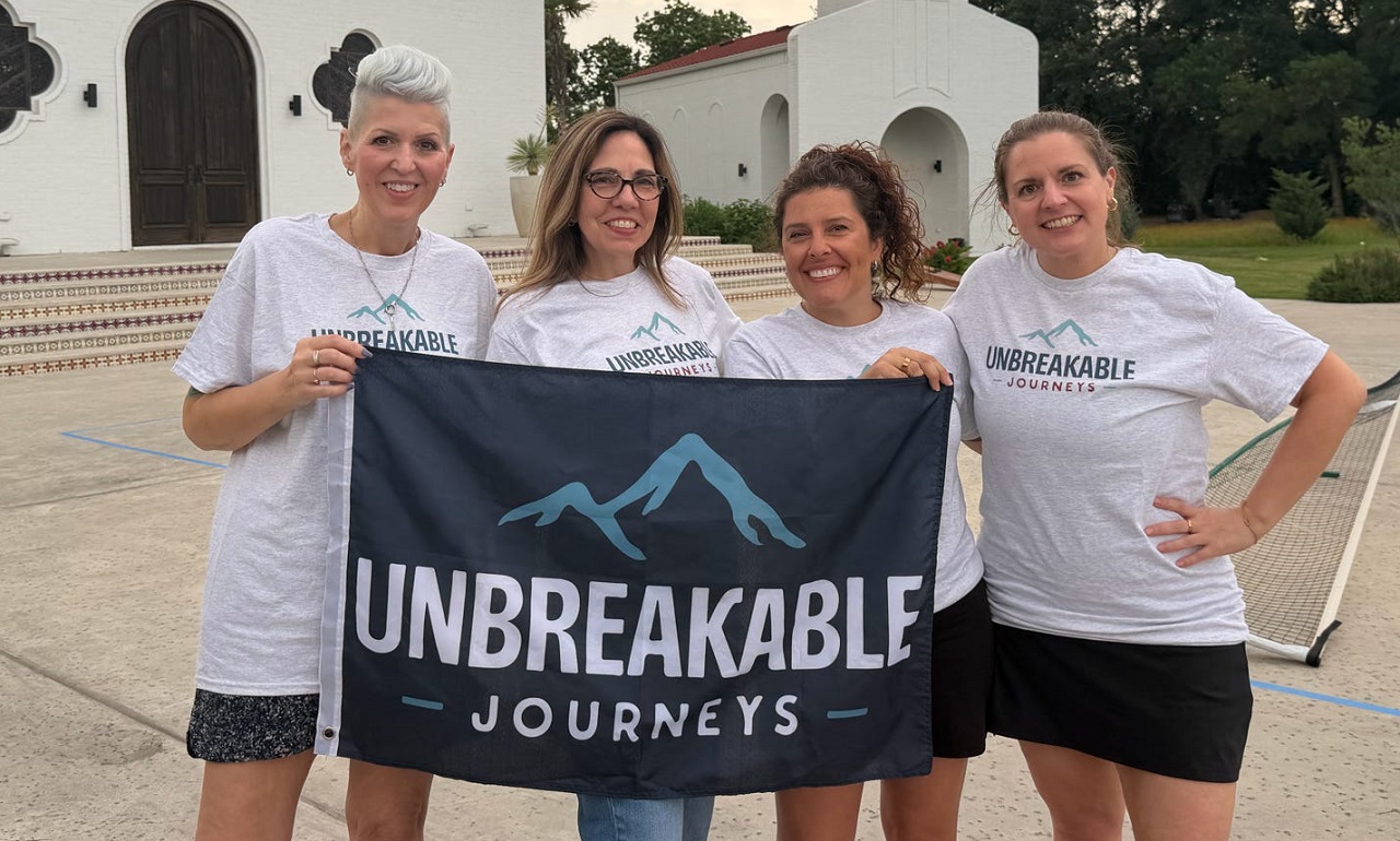 Photo May 17 2026, 6 13 50 PM Four women stand outdoors, smiling and holding a banner that reads Unbreakable Journeys. All are wearing matching Unbreakable Journeys T-shirts. A white building with arched doors is in the background.