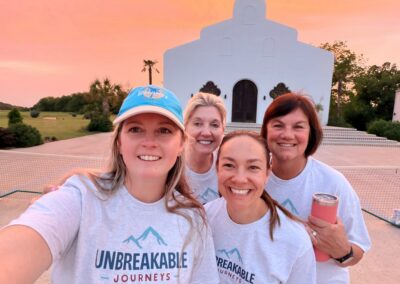 Four women in matching Unbreakable Journeys shirts smile for a selfie outside a white building at sunset, with trees and a net visible in the background.