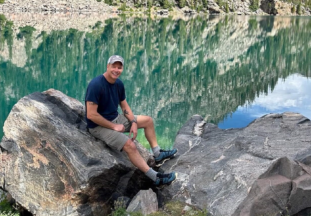 shanehero A man in a cap and shorts sits smiling on large rocks by a clear, reflective lake with mountains and trees in the background.
