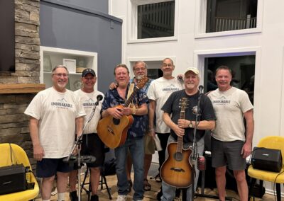 Seven smiling men stand indoors, some holding guitars, with microphones and music equipment around. Four men wear matching UNBREAKABLE shirts; the atmosphere is casual and friendly, suggesting a musical gathering or band practice.