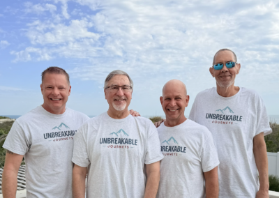 Four smiling men in Unbreakable Journeys shirts stand outdoors near a beach, with blue sky and scattered clouds in the background.
