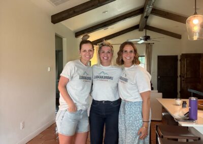Three women stand and smile in a kitchen, each wearing matching gray shirts. They stand close together with arms around each other, in a room with wooden ceiling beams and terra-cotta tile floor.