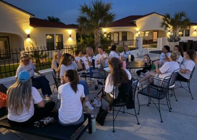 A group of women in white shirts sit around tables outdoors in the evening, talking and laughing. Palm trees and well-lit buildings are in the background.