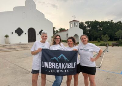 Four women stand in front of a white mission-style building holding a flag that reads UNBREAKABLE JOURNEYS with a mountain icon.