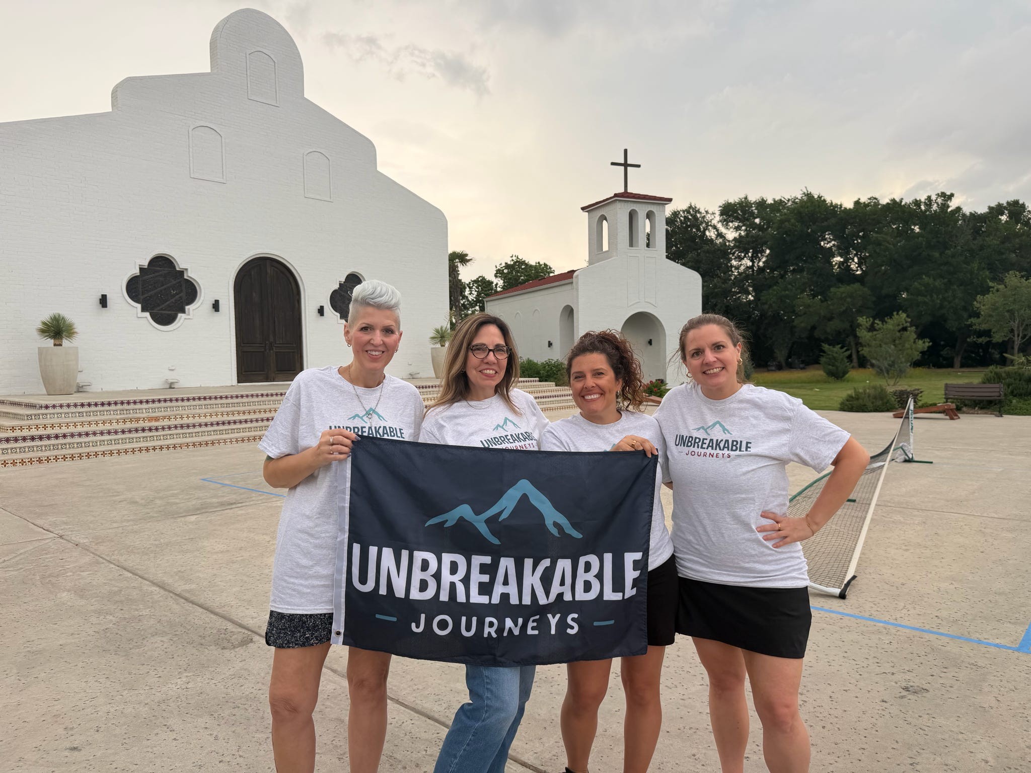 Four women stand in front of a white mission-style building holding a flag that reads UNBREAKABLE JOURNEYS with a mountain icon.