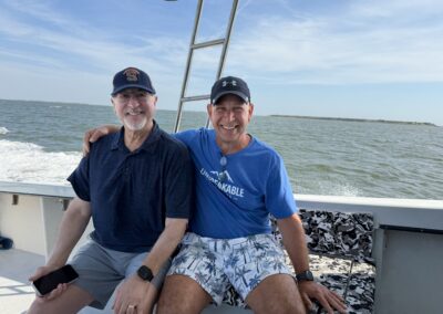 Two men in baseball caps sit side-by-side on a boat with the ocean in the background.