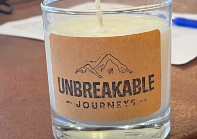 A glass jar candle with a kraft paper label reading UNBREAKABLE JOURNEYS under a mountain line drawing.