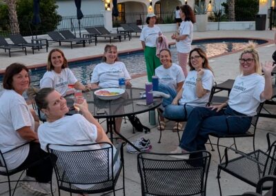 A group of women in matching white t-shirts sit around a patio table by a swimming pool at sunset.