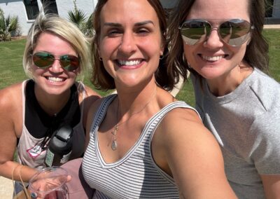 Three women smiling together outdoors in front of a white house with a red roof.