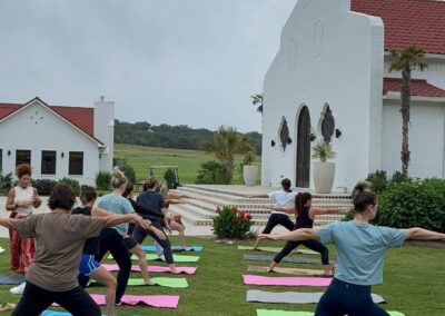 A group of people perform yoga poses on colorful mats in a grassy lawn in front of a white building.