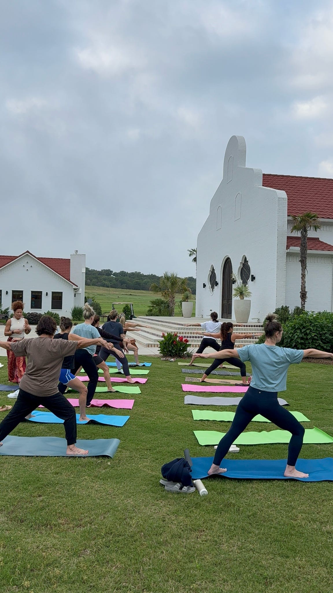 A group of people perform yoga poses on colorful mats in a grassy lawn in front of a white building.